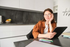 Eine junge Frau mit Brille und brauner Bluse sitzt in einer modernen Küche am Tisch, hält ein Tablet und blickt nachdenklich zur Seite.