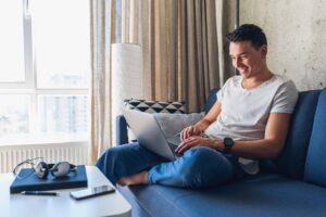 Ein junger Mann sitzt mit einem geöffneten Laptop auf einem dunkelblauen Sofa neben einem großen Fenster.