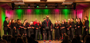 Der Chor Edentity steht in schwarz-roter Kleidung auf einer bunt beleuchteten Bühne und singt vor Publikum. Im Vordergrund dirigiert der Chorleiter mit dem Rücken zur Kamera.