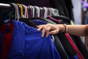 Eine Hand greift nach einer blauen Jacke an einer Kleiderstange mit zahlreichen bunten Kleiderbügeln.