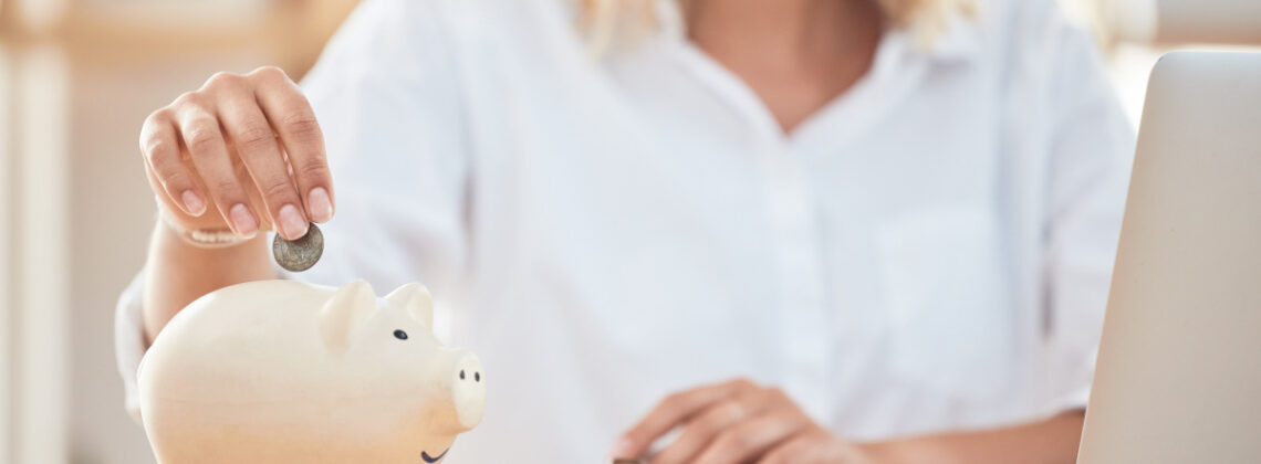 Eine blonde Frau, die eine Münze in ein Sparschwein schmeißt und daneben steht ein geöffneter Laptop, Symbolbild für Frauen und Finanzen