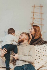 Eine Mutter und ein Vater sitzen mit ihrem Kleinkind auf einem hellen Bett. Der Vater hält das Kind auf dem Arm, während die Mutter lächelnd zusieht.