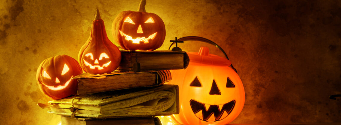 Mehrere leuchtende Halloween-Kürbisse stehen neben und auf einem Stapel alter Bücher vor dunkel-orange beleuchtetem Hintergrund.
