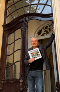 Porträt Frank Brünnee: Lächelnder Mann steht in einem kunstvoll verzierten historischen Hauseingang, hält das Buch „Digitale Kunstgeschichte“ in der Hand und stützt sich mit dem anderen Arm am Türrahmen ab.